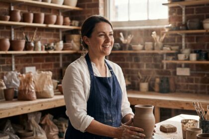 Artisane en tablier de lin souriante dans son atelier de céramique, mains posées sur une pièce en argile, ambiance chaleureuse et authentique